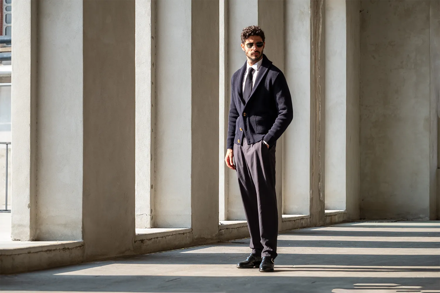 Un uomo in piedi con cardigan blu, cravatta e pantaloni eleganti, ripreso in un piazzale di un'area industriale