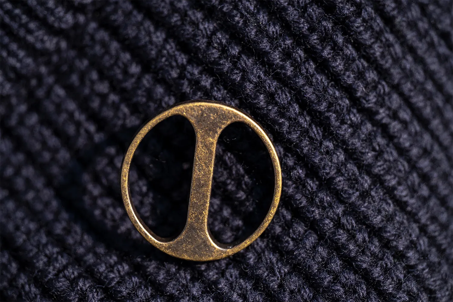 Primo piano di un bottone in stile vintage dorato, rappresentante il logo del brand, fissato su un maglione di lana blu scuro