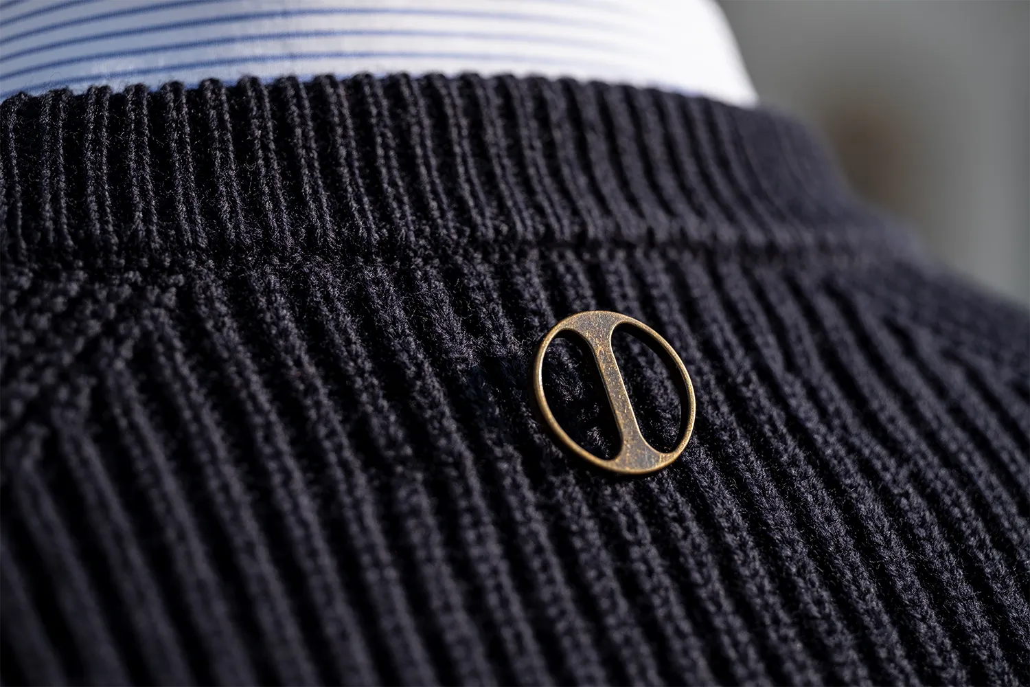 Dettaglio di un bottone dorato anticato con logo del brand, posizionato al centro delle spalle sotto il collo su un maglione di lana blu
